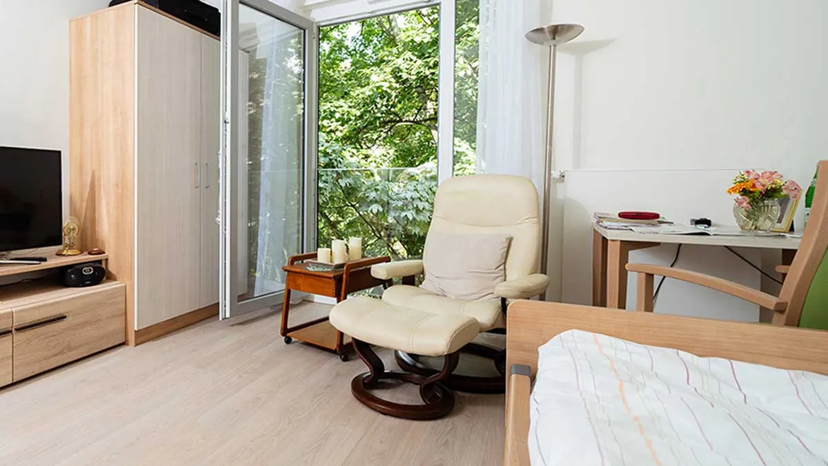 Zimmer im PFLEGEN & WOHNEN HAMBURG-Standort Marienthal mit Sessel, Bett, Schreibtisch mit Stuhl, Schrank und einem Fernseher.